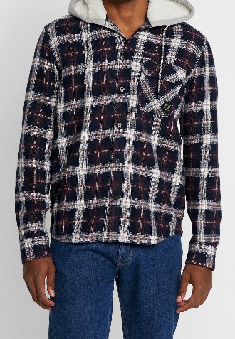 Plaid flannel shirt in navy, red, and white, featuring a soft grey-lined hood, button-down front, and a chest pocket with patch details.