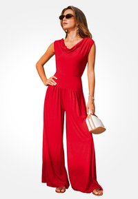 Mono rojo sin mangas con cuello drapeado, diseño de pierna ancha y cintura elástica. Combinado con gafas de sol y un pequeño bolso beige.