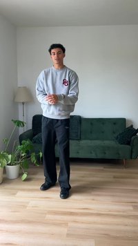 Šedá mikina s bordó logem, černé kalhoty a černé boty. Stojí na dřevěné podlaze blízko zelené pohovky a květiny v květináči. Minimalistická výzdoba.