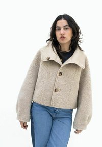 Beige sherpa jack met een oversized kraag, knoopsluiting en wijde mouwen. De stof heeft een gestructureerde, gezellige uitstraling. Gecombineerd met blauwe jeans.