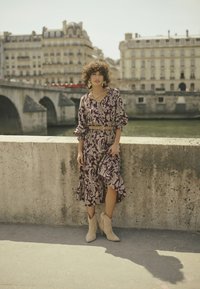 Lila mit Blumenmuster versehendes Kleid mit Puffärmeln und seitlichem Schlitz, akzentuiert durch einen beigen Gürtel. Trägt beige Stiefeletten mit Absatz.