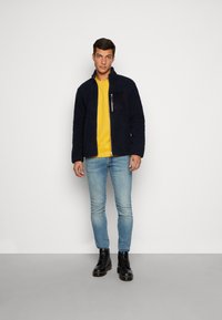 Donker marineblauwe fleece jas met een ritssluiting op de borst, gecombineerd met een felgeel shirt, lichtblauwe skinny jeans en zwarte veterschoenen.