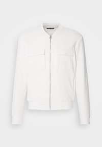 Chaqueta bomber blanca con una tela texturizada, cierre frontal con cremallera, dos bolsillos en el pecho y puños y dobladillo de canalé. Diseño minimalista.
