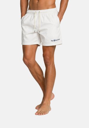 Hombre que lleva pantalones cortos de baño blancos con cordón y el logotipo de Polo Ralph Lauren en la pierna izquierda, de pie descalzo con las manos en los bolsillos.