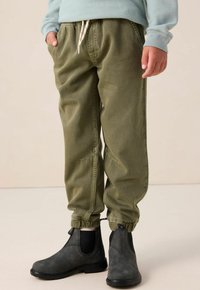 Olijfgroene joggerbroeken met een elastische tailleband en trekkoord, voorzien van zijzakken en elastische boorden. Gecombineerd met zwarte enkellaarsjes.