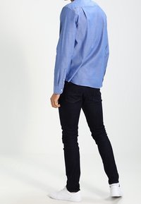 Jasnoniebieska, długa koszula z guzikami i kołnierzykiem, w połączeniu z ciemnymi dżinsami i białymi trampkami. Gładka tekstura, dopasowany krój.