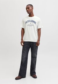 Hvid bomuldst-shirt med blå grafisk tekst, kombineret med mørkegrå vidde-bukser og brune sko. Stoffet virker blødt med en afslappet pasform.