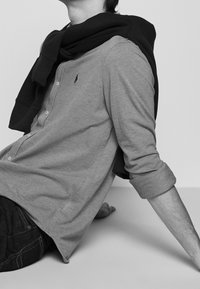 Šedá košile s dlouhým rukávem a knoflíky, na krku přehozená černá mikina. Na hrudi je vyšitý logo.