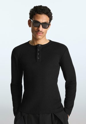 Junger Mann trägt ein schwarzes, geripptes Langarmshirt mit Knöpfen, dunkle Sonnenbrille und dunkle Hose, steht vor einem einfarbigen hellen Hintergrund.