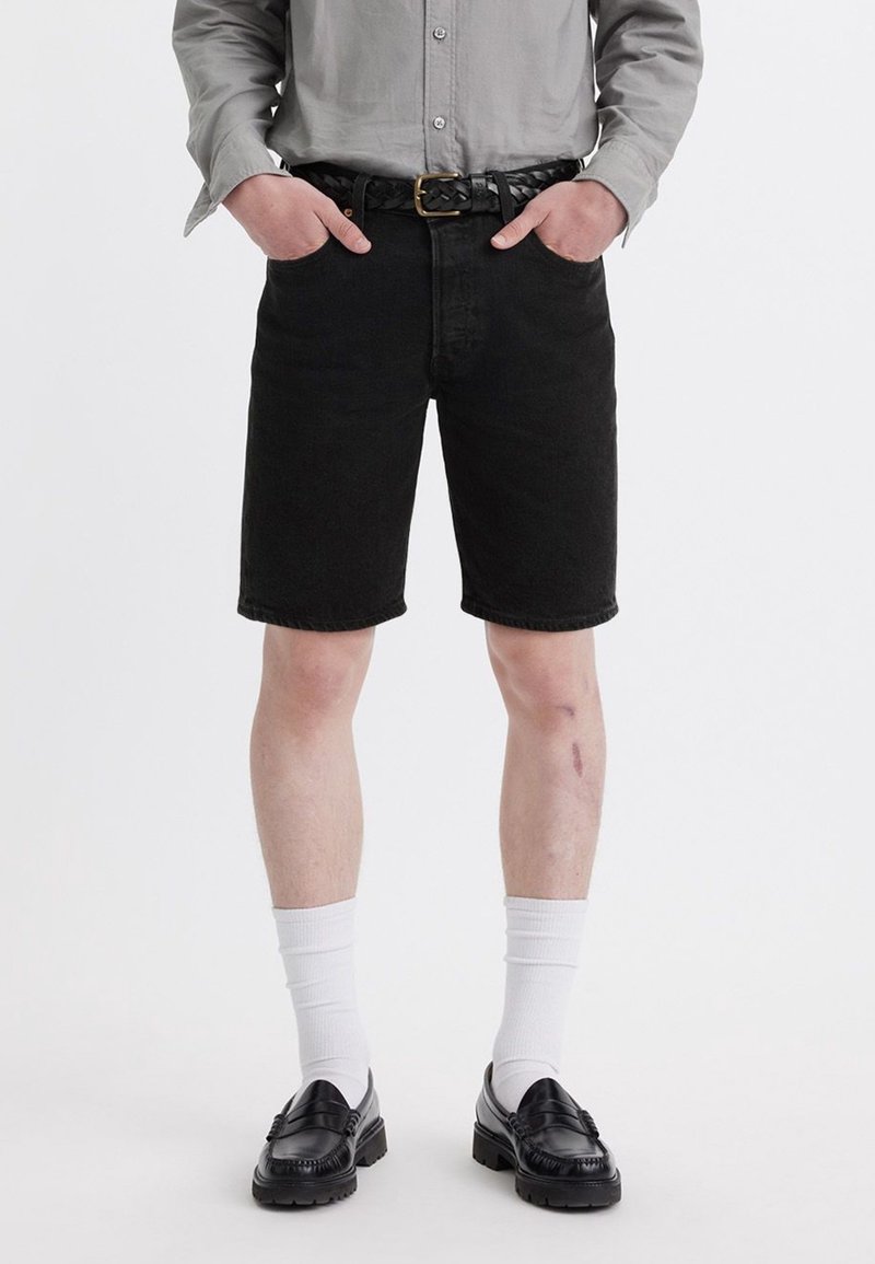 Homme portant un short en jean noir, ceinture noire, chemise grise à manches longues, chaussettes blanches et chaussures noires, debout les mains dans les poches.