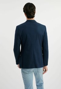 Granatowa marynarka dopasowana, wykonana z gładkiego materiału, z pojedynczym wentylem i zapięciem na dwa guziki, noszona na jasnoniebieskich dżinsach.