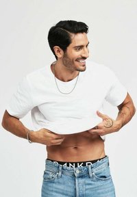 Vit kortärmad T-shirt upplyft för att visa det svarta märkeskantbandet på jeansen. Modellen har mörkt hår och synliga tatueringar på armarna.