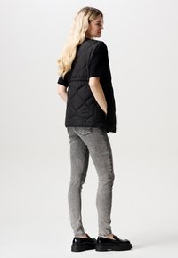 Svart kviltad väst med fickor, bärs över en svart topp, kombinerad med grå smala jeans och svarta plattformsskor. Mjuk textur och slät yta.