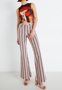 Femme portant un haut sans manches imprimé flammes de match, un pantalon évasé à taille haute rayé, et des chaussures à talons blocs ouvertes couleur nude sur un fond blanc.