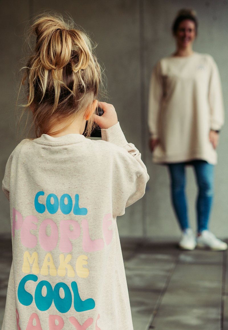 Hellbeige Sweatshirt mit buntem Text "COOL PEOPLE MAKE COOL BABIES" auf dem Rücken. Ein Kind hält eine Kamera und schaut weg.