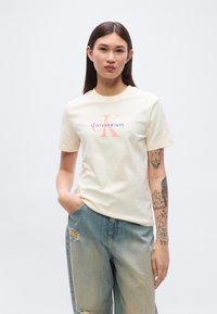 Μπλουζάκι από βαμβάκι σε κρ εμ απόχρωση με ένα λογότυπο σε ροζ και μοβ, συνδυασμένο με ελαφρώς ξεβαμμένα τζιν με φθορές. Η εφαρμογή είναι άνετη και χαλαρή.