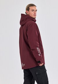 Maroon hooded jacket with a relaxed fit, featuring a logo on the sleeve and a black accent near the wrist. Worn over black cargo pants.