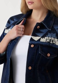 Džinsinė striukė su mėlynos spalvos raštuota mezgimo apykakle, variniais mygtukais ir struktūrine apykakle. Sukurkta su mygtukų uždarymu ir šoninėmis kišenėmis.