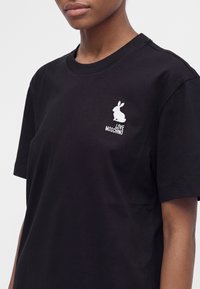 Άτομο που φοράει μαύρο t-shirt Moschino με μικρό λευκό κεντητό κουνέλι και το κείμενο "LOVE MOSCHINO" στο στήθος.