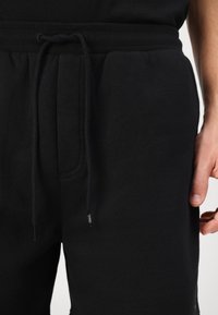 Schwarze Jogginghose mit elastischem Bund und Kordelzug, Nahaufnahme der Taille und der rechten Hand einer Person, die neben dem Bein ruht.