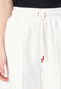 Pantalones jogger blancos con una cinturilla acanalada, que incluyen cordones con acentos rojos. Tejido suave y diseño con pliegues para un detalle adicional.