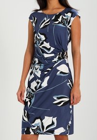 Mujer con un vestido azul de longitud justo por la rodilla y mangas cortas, con grandes patrones florales en blanco, negro y azul claro, y recogidos en los laterales.