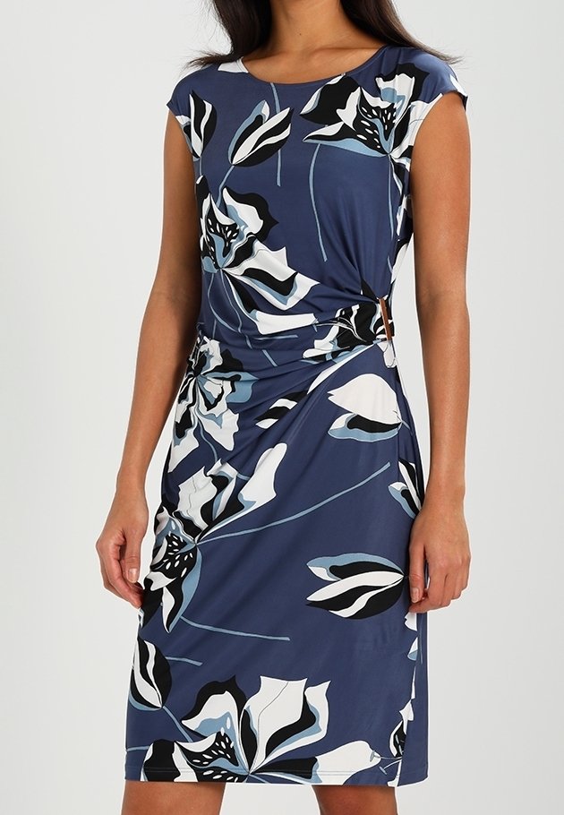 Mujer con un vestido azul de longitud justo por la rodilla y mangas cortas, con grandes patrones florales en blanco, negro y azul claro, y recogidos en los laterales.