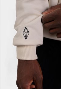 Manica di felpa crema con polsino a coste; presenta un logo a diamante ricamato in nero. La mano che afferra il tessuto mostra texture e contrasto di colore.