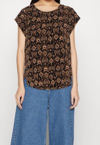 Svart blus med ett mönstrat design i nyanser av brunt och beige. Har korta ärmar och rundad halsringning. Kombineras med vida denimbyxor.