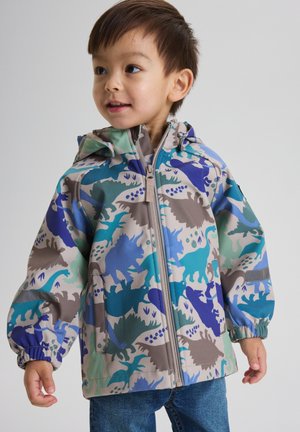 Junger Junge trägt eine bunte Kapuzenjacke mit Dinosaurier-Motiv und blaue Jeans, schaut seitlich vor einem schlichten Hintergrund.
