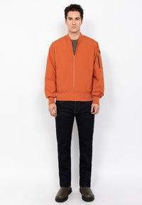 Junger Mann trägt eine orange Zip-Up Bomberjacke, dunkle Jeans und olivgrüne Stiefel und steht vor einem schlichten weißen Hintergrund.