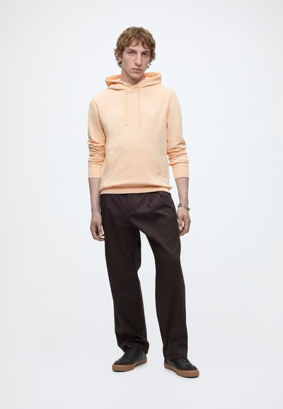 Homme portant un sweat à capuche pêche et un pantalon marron foncé, debout les mains détendues, sur un fond blanc uni.