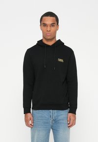Joven con una sudadera negra con capucha y bolsillo frontal, con el logo dorado de "Karl Lagerfeld", combinada con jeans azul claro, de pie contra un fondo blanco.
