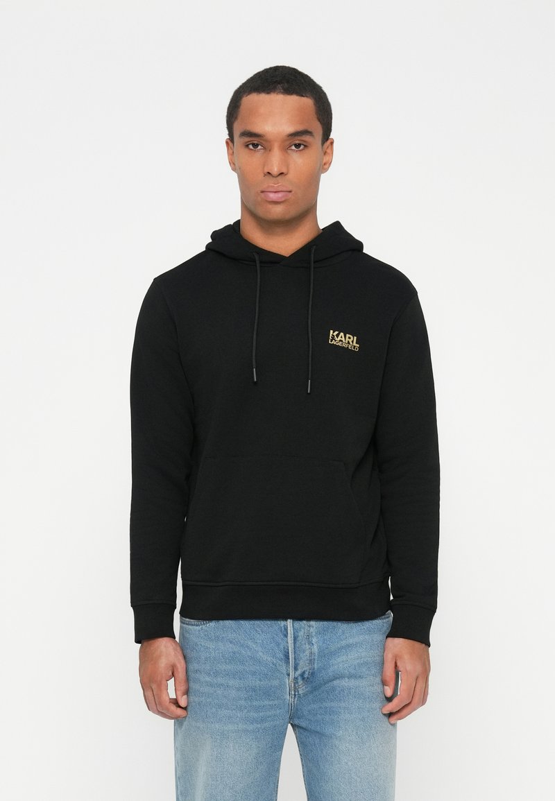 Joven con una sudadera negra con capucha y bolsillo frontal, con el logo dorado de "Karl Lagerfeld", combinada con jeans azul claro, de pie contra un fondo blanco.