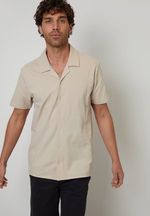 Muž s kudrnatými vlasy, který má na sobě béžovou košili s krátkým rukávem na knoflíky a černé kalhoty na jednoduchém světlém pozadí.