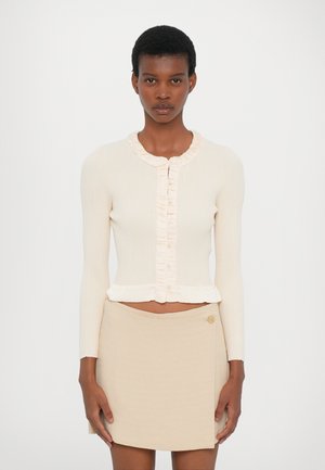 Kremowy kardigan prążkowany z falbaniastym zapięciem i mankietami, w połączeniu z beżową spódnicą kopertową z detalem guzika. Krótki, dopasowany fason.