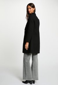 Sort uldblandingsfrakke med skræddersyet pasform, notched krave, to forlommer og gyldne knapper, parret med bredlårede grå jeans.