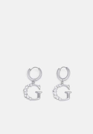 Boucles d'oreilles créoles en argent avec des breloques en forme de lettre G suspendues, partiellement serties de petites pierres précieuses transparentes sur un fond blanc uni.