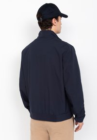 Veste légère bleu marine avec un col haut, un ourlet élastique et des poignets boutonnés. Texture lisse sans motifs visibles.