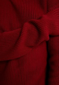 Maglione rosso lavorato a maglia con un design a costine testurizzato, maniche arrotolate e un'aspetto morbido e caldo.