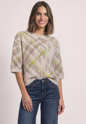 Femme aux cheveux bruns mi-longs, portant un pull beige à manches courtes à motifs et un jean bleu, souriante, debout devant un fond uni.