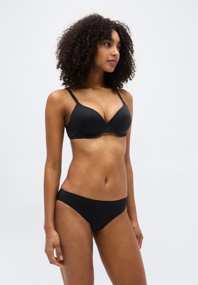 Γυναίκα με σγουρά μαλλιά που φορά μαύρο σουτιέν και μαύρο εσώρουχο, στέκεται μπροστά σε ένα απλό ανοιχτόχρωμο φόντο, κοιτώντας ελαφρώς προς τα δεξιά.