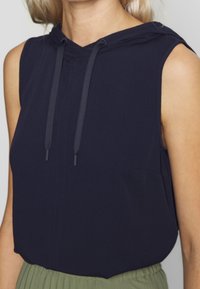 Haut sans manches marine avec capuche, doté de cordons de serrage, d'une coupe décontractée et d'une poche avant, associé à un bas plissé vert clair.