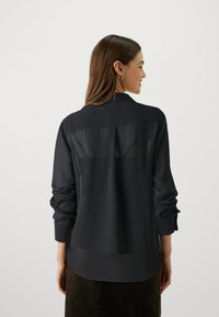 Black, sheer blouse with long sleeves and a rounded hem. Features a collar and subtle back pleat detail, made from lightweight fabric.