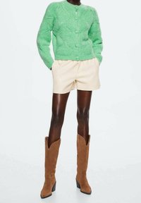 Osoba s tmavou pletí oblečená v zeleném pleteném kardiganu, béžových kraťasech a hnědých semišových kozačkách až po kolena, stojící s rukama v kapsách.