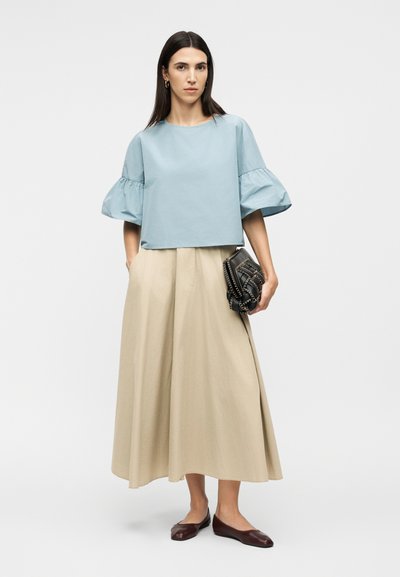 Γυναίκα που φοράει γαλάζιο πουκάμισο με φουσκωτά μανίκια, μπεζ φαρδιά φούστα, κρατάει μαύρο καπιτονέ τσαντάκι και φοράει καφέ φλατ παπούτσια μπροστά σε μονόχρωμο φόντο.