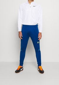 Bílé dlouhé rukávy s čtvrtinovým zipem a černým logem; modré sportovní kalhoty se žlutým logem; černé a oranžové fotbalové kopačky s bílými akcenty.