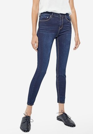 Dunkelblaue Skinny-Jeans aus Denim mit mittelhohem Bund, fünf Taschen, kontrastierenden Nähten und einer glatten Textur. Kombiniert mit schwarzen Schuhen.