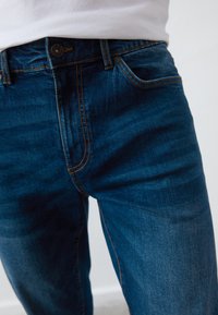 Tumši zili denim džinsi ar oranžiem izšuvumiem, ar pogu aizdari, jostu cilpām un gludu tekstūru. Tuvplāns no priekšpuses.