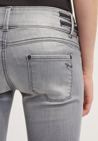 Jeans de mezclilla gris con un diseño ajustado, que presentan un bolsillo trasero con un logo sutil y costuras decorativas. Detalle de cremallera en la cintura.